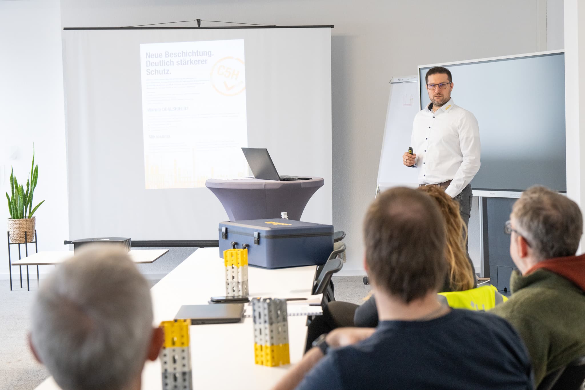 Man in white shirt giving a presentation to seated attendees in a conference room with a projector screen, laptop and flip chart.