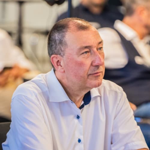 Man in a white shirt seated and attentively listening in an indoor setting with blurred individuals in the background.