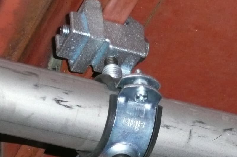 Metal clamp securing a cylindrical pipe to a red surface with bolts, showing close-up of industrial fastening components.