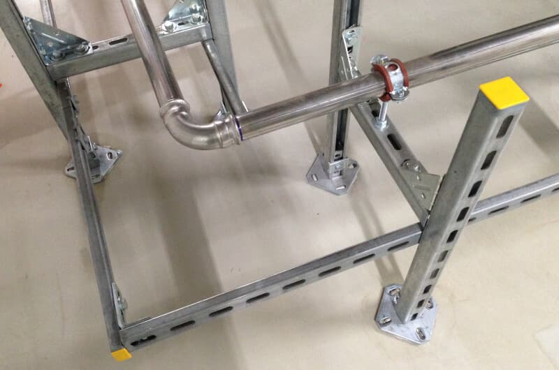 Steel framework with bolted connections, featuring horizontal and vertical beams. A metal pipe secured by a bracket runs alongside.