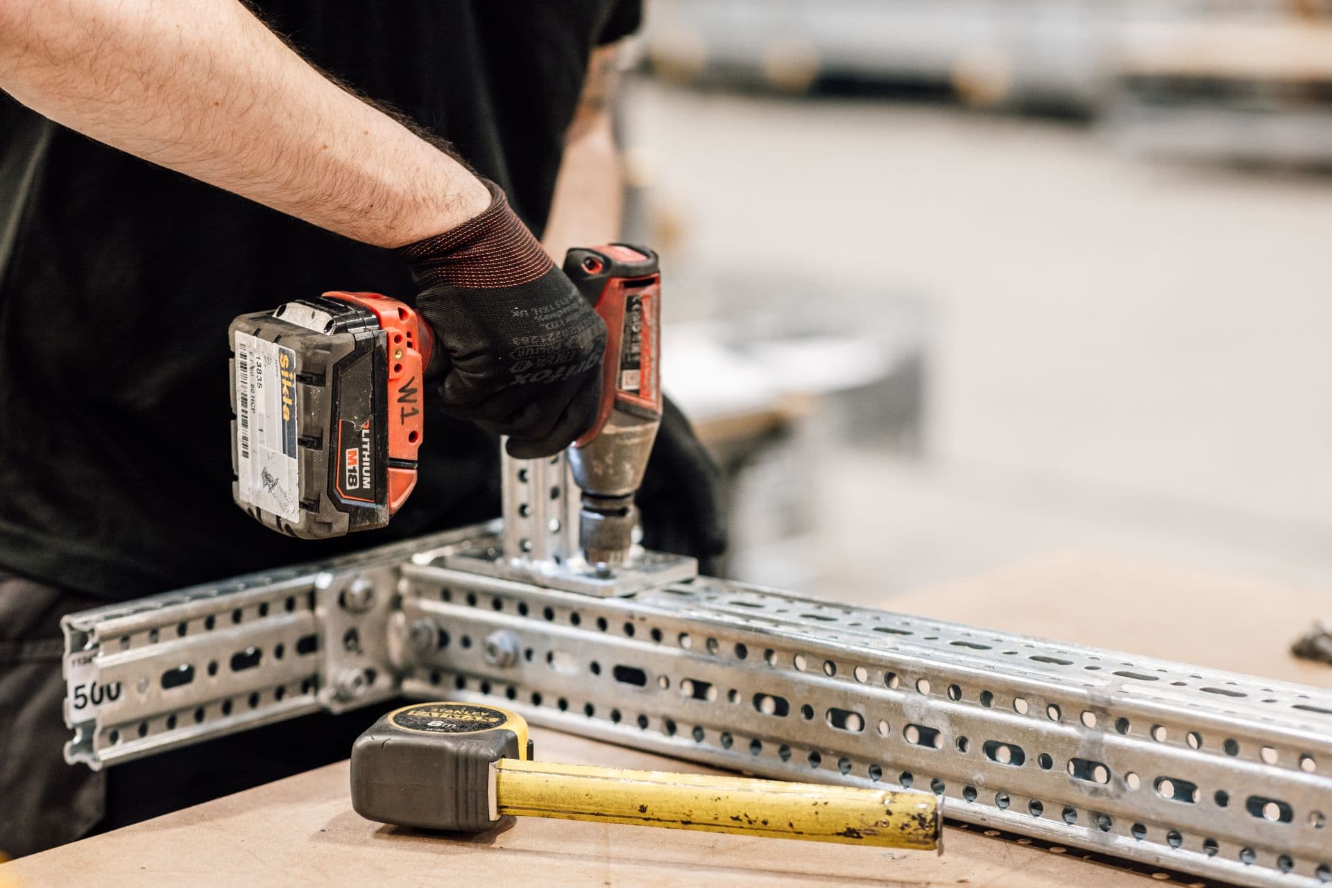 Person using a power drill on steel beams, with a yellow tape measure nearby on a workbench.