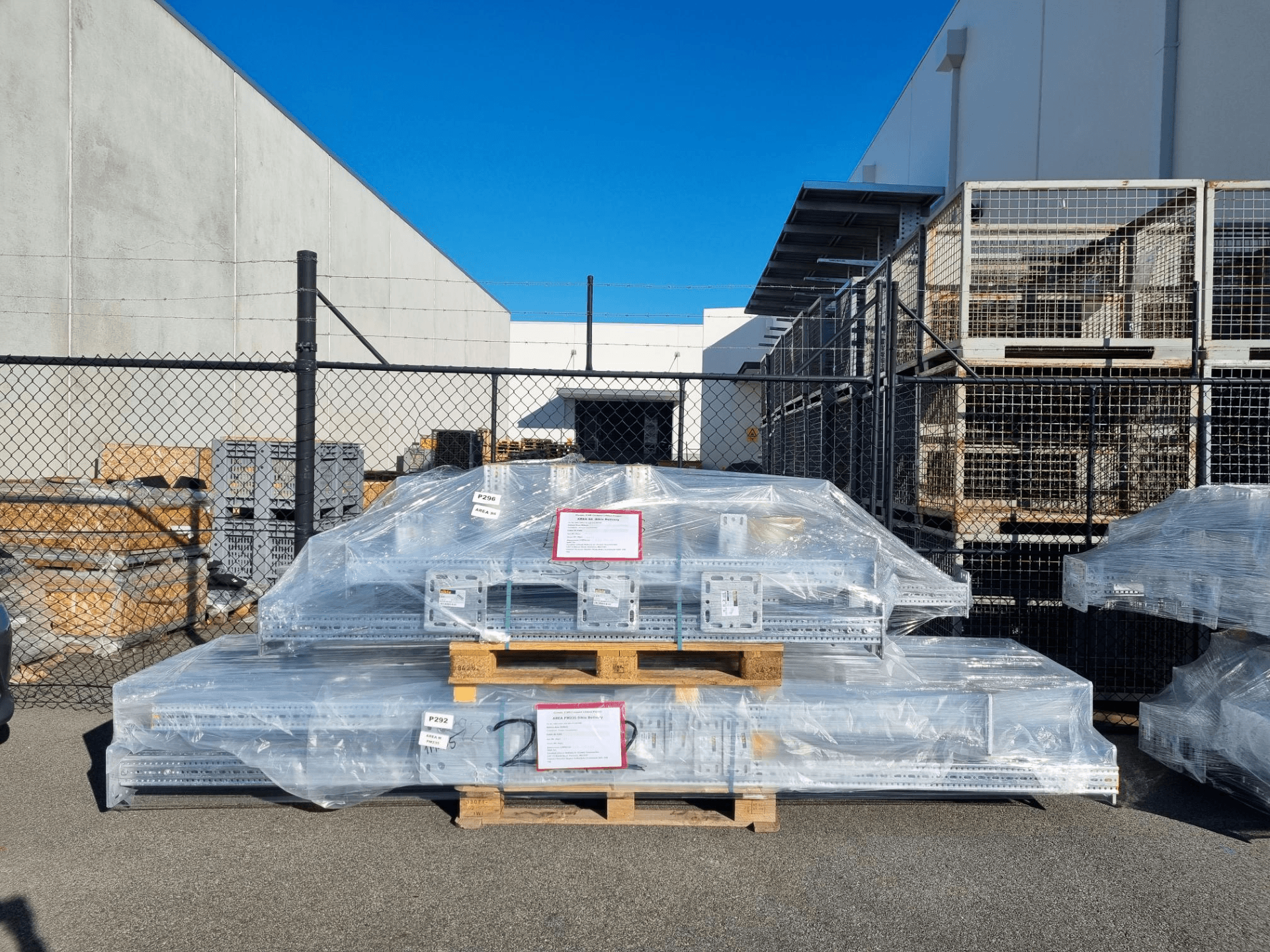 Piles of metal construction materials wrapped in plastic are stacked on wooden pallets outside, surrounded by a metal fence under a clear blue sky.