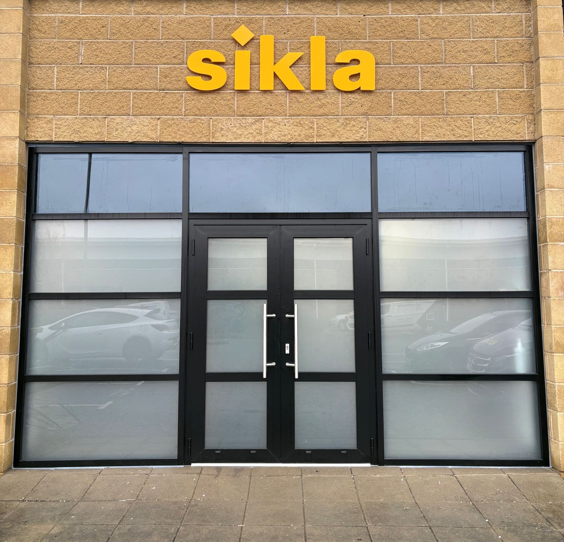 A office front with black-framed glass doors and windows, featuring a yellow "sikla" sign above.